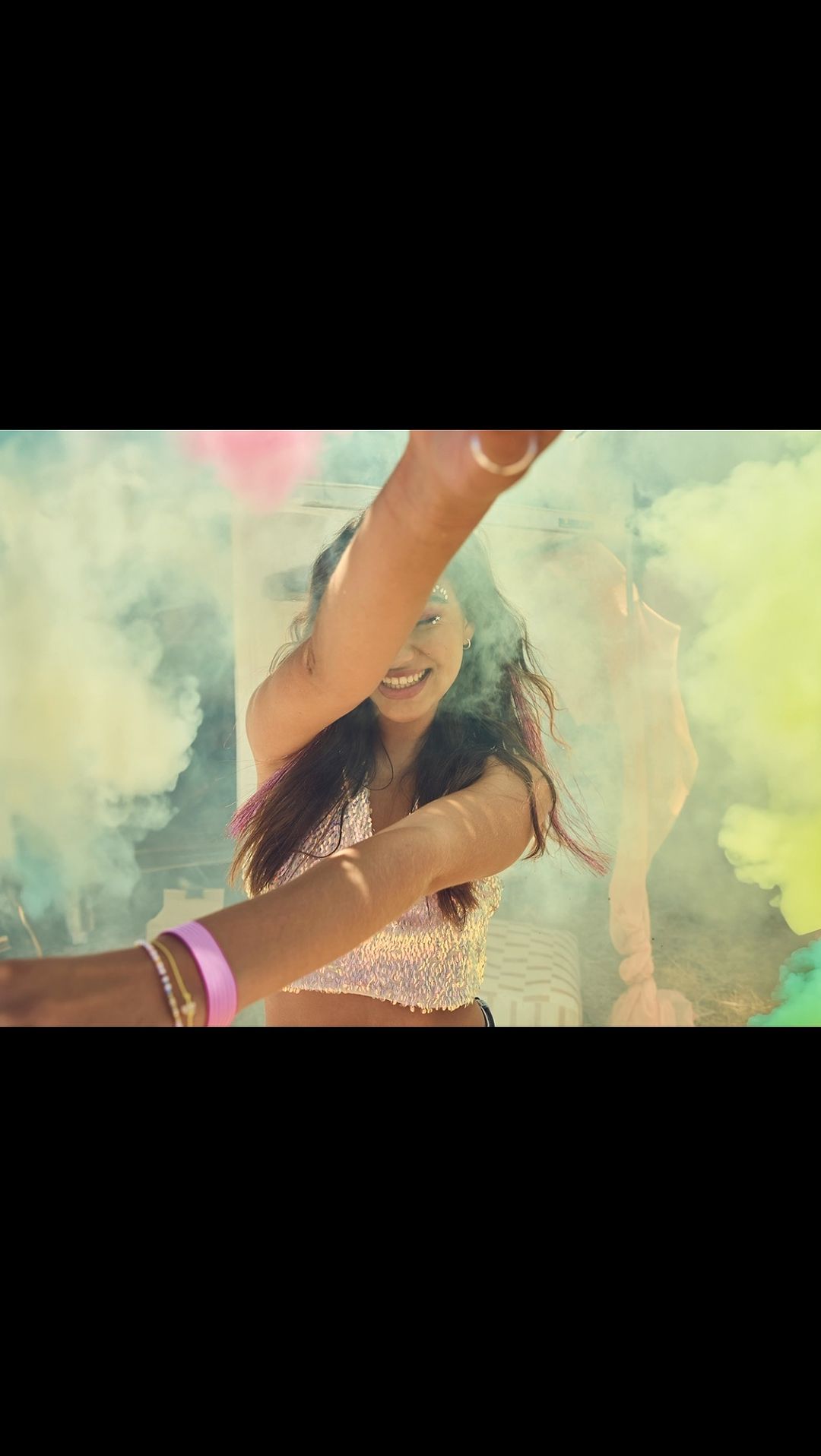 Smiling woman with arms outstretched poses amid vibrant smoke clouds in an outdoor setting, wearing a glittery top, and bracelets.