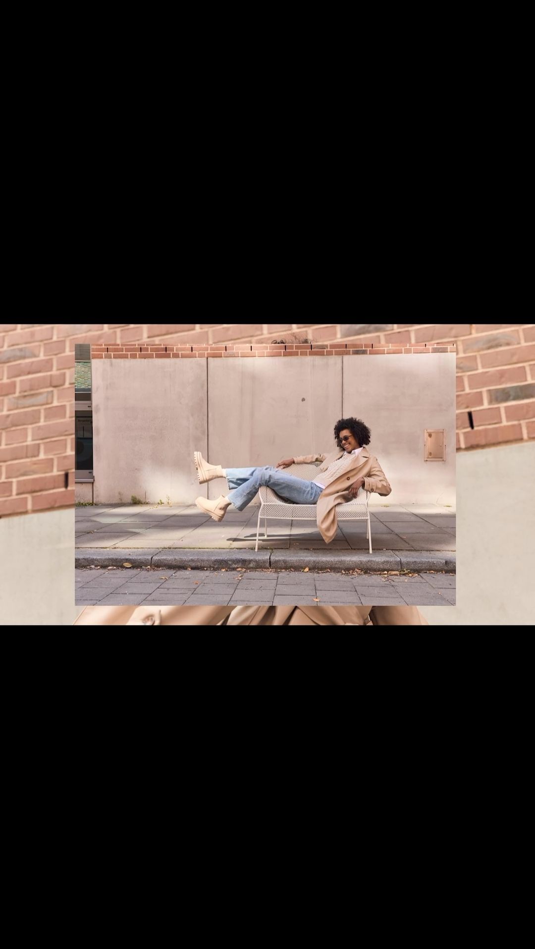 Woman in sunglasses poses on a chair outdoors in blue jeans, a tan blazer and cream boots. Brown brick and pavement visible.