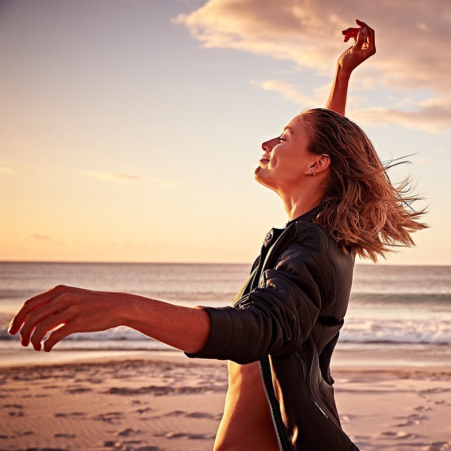 Blonde woman stands on a beach with arms outstretched at sunset. She is wearing a black jacket, and the sun reflects off the ocean.