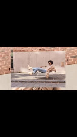 Black woman with curly hair wearing a tan coat and blue jeans relaxing on a white metal chair on a paved sidewalk.