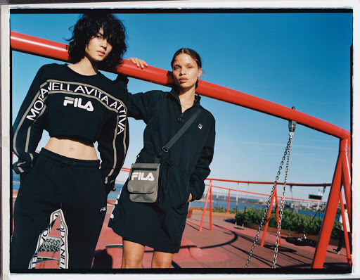 Two young women in Fila activewear pose outdoors against a bright blue sky, red architectural structure, and distant cityscape.