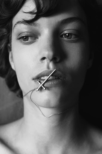 Close-up portrait of a woman holding a silver cross pendant necklace in her mouth with make-up by Laure Dansou.