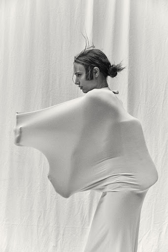 Monochrome image of a person in a full-body garment standing against a draped white backdrop, creating a minimalist and abstract composition.