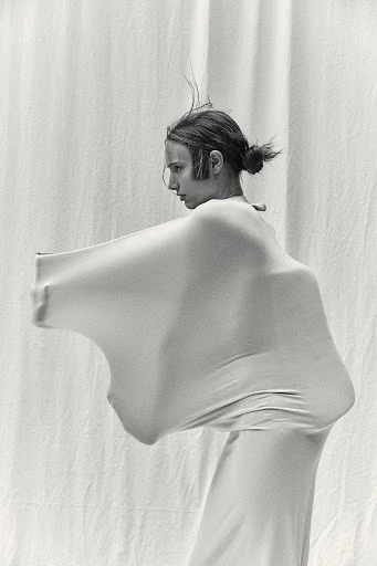 Monochrome image of a person in a full-body garment standing against a draped white backdrop, creating a minimalist and abstract composition.