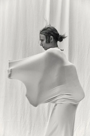 Monochrome image of a person in a full-body garment standing against a draped white backdrop, creating a minimalist and abstract composition.