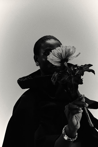 Black and white portrait of a black woman holding a flower in front of her face wearing a dark coat and jewelry, with soft lighting.