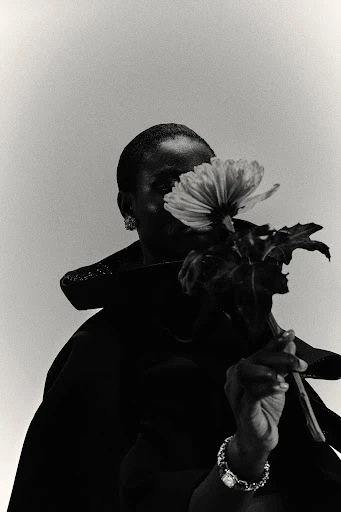 Black and white portrait of a black woman holding a flower in front of her face wearing a dark coat and jewelry, with soft lighting.