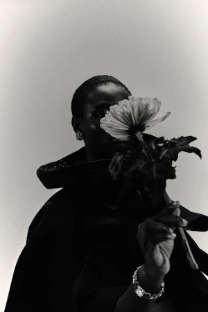 Black and white portrait of a black woman holding a flower in front of her face wearing a dark coat and jewelry, with soft lighting.