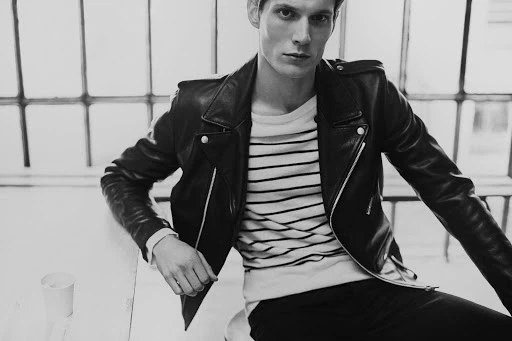 Monochrome portrait of a handsome male model in a leather jacket and striped shirt, sitting indoors near a window with natural lighting.