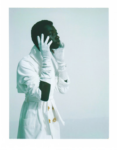 Elegant portrait of a Black male in a white coat and gloves against a bright background, showcasing a modern, fashion-forward aesthetic.