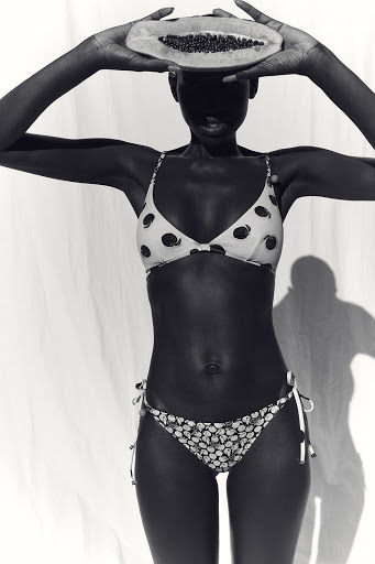Black and white photo of a slender dark-skinned woman in a patterned bikini holding a halved papaya fruit over her face in a studio.