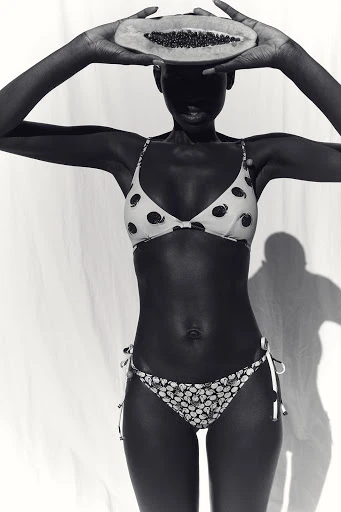 Black and white photo of a slender dark-skinned woman in a patterned bikini holding a halved papaya fruit over her face in a studio.