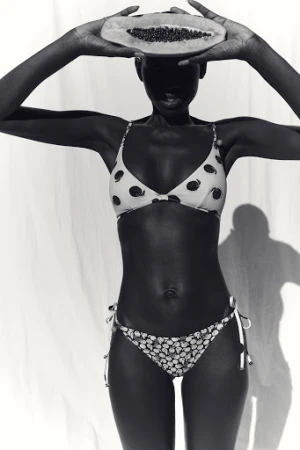 Black and white photo of a slender dark-skinned woman in a patterned bikini holding a halved papaya fruit over her face in a studio.