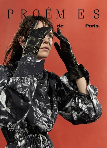 Fashion shot of a woman in a patterned dress and black gloves, posing with her hands up against a terracotta background with text.