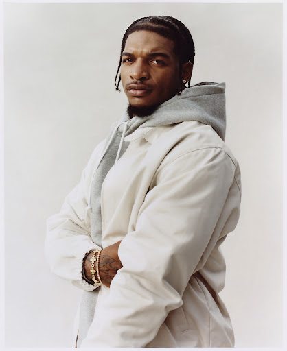 Studio portrait of a Black male model in a neutral hoodie and jacket with gold bracelets, set against a clean, bright background.