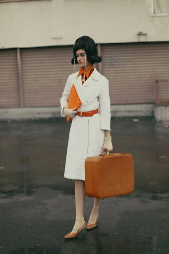 A woman with a beehive hairdo in a vintage coat, stylish glasses, and heels, holding a briefcase and suitcase against a gray backdrop.