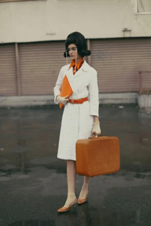 A woman with a beehive hairdo in a vintage coat, stylish glasses, and heels, holding a briefcase and suitcase against a gray backdrop.
