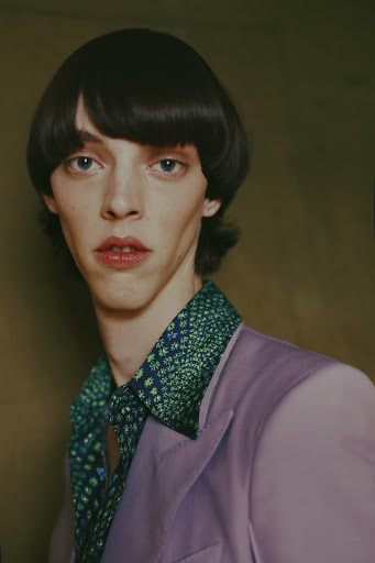 Portrait of a person with dark bowl-cut hair wearing a purple jacket and patterned green collared shirt against a dark background.