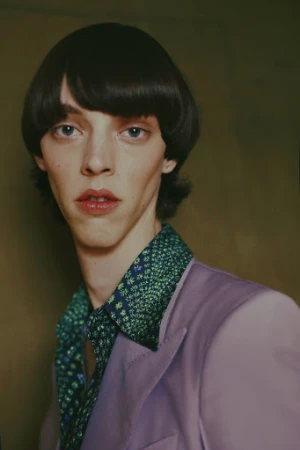 Portrait of a person with dark bowl-cut hair wearing a purple jacket and patterned green collared shirt against a dark background.