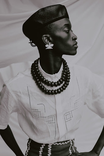 Portrait of a dark-skinned person wearing a hat, beaded necklace, and patterned shirt, facing right against a neutral backdrop in a studio.