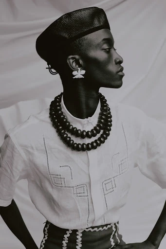 Portrait of a dark-skinned person wearing a hat, beaded necklace, and patterned shirt, facing right against a neutral backdrop in a studio.