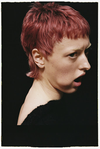 Close-up profile of a person with short pink textured hair and dark clothing against a black background with a white polaroid border