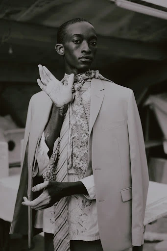 Black and white portrait of a slender, dark-skinned man in a blazer and floral tie, hand raised, with a background of blurred shapes.