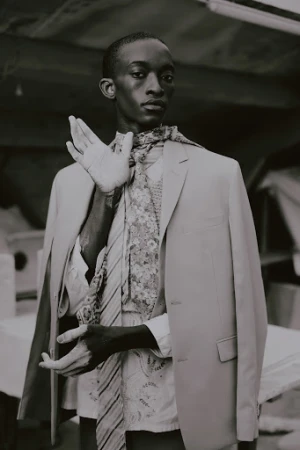 Black man in tailored blazer and patterned scarf posing in a dimly lit industrial interior space.