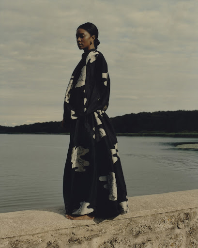 A lone adult female stands on a stone ledge near calm water under a cloudy sky, wearing a flowing black and white patterned dress.