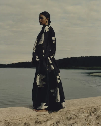A lone adult female stands on a stone ledge near calm water under a cloudy sky, wearing a flowing black and white patterned dress.