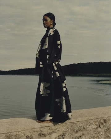 A lone adult female stands on a stone ledge near calm water under a cloudy sky, wearing a flowing black and white patterned dress.