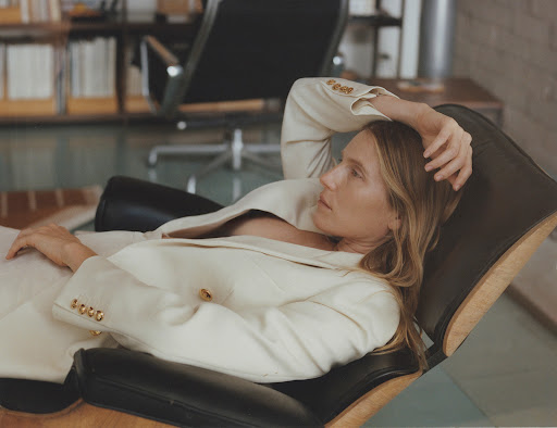 Woman in a white suit reclines in a black leather chair in an indoor setting, with natural light illuminating the scene.
