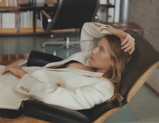 Woman in a white suit reclines in a black leather chair in an indoor setting, with natural light illuminating the scene.