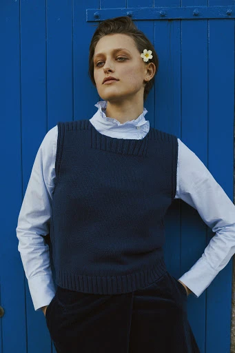 A woman with short hair in a blue sweater vest over a white blouse poses in front of a matching blue wooden door for a fashion shot.