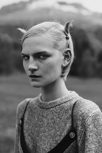 Monochrome portrait of a blonde woman with styled hair, wearing a grey sweater with black straps, set against a blurred mountain backdrop.