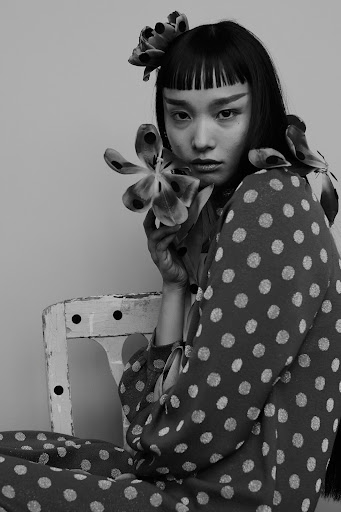 Portrait of a young Asian woman with unique eye makeup, polka dot dress, and flowers in her hair, seated against a neutral backdrop.