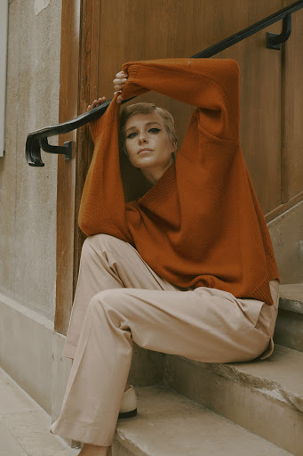 A fair-skinned woman with short blonde hair sits on stone steps, wearing an orange sweater and light pink pants in a lifestyle setting.