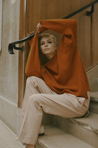 A fair-skinned woman with short blonde hair sits on stone steps, wearing an orange sweater and light pink pants in a lifestyle setting.
