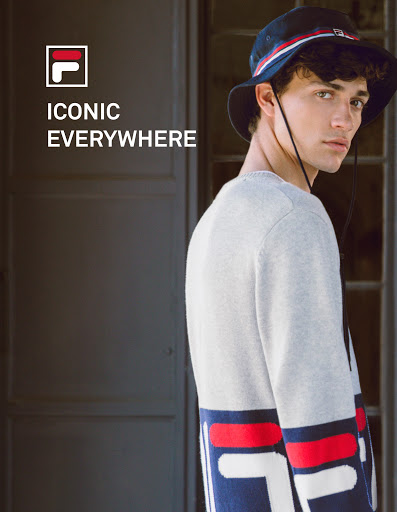 Young man wearing a navy bucket hat and grey sweater with red and white branding, looking over his shoulder.