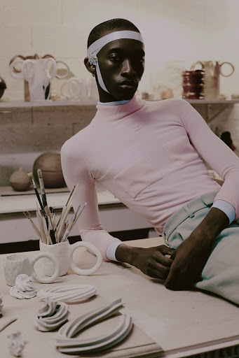 Young adult poses in a pink turtleneck with ceramic art objects on a table in a bright studio, showcasing a blend of fashion and craft.