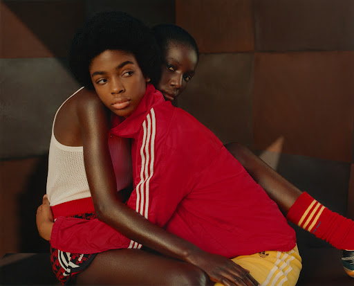 Two models in athletic wear embrace against a geometric backdrop, featuring one in a white top and another in a red track jacket.