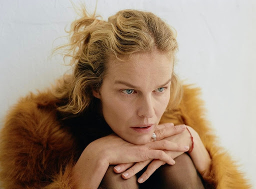 Portrait of adult female with blonde hair wearing a brown faux fur coat, seated indoors against a white wall in soft, diffused light.