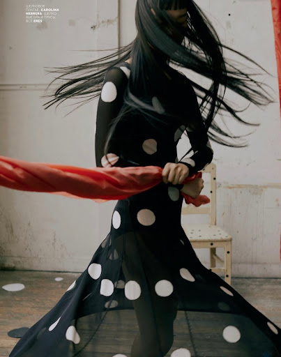 Model in a black and white polka dot dress with long flying hair holding a red fabric banner.