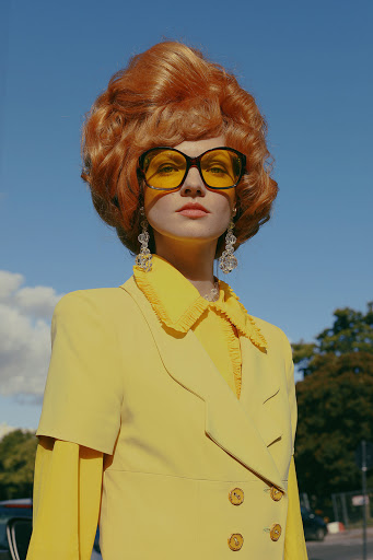 Stylish woman with large auburn hair and sunglasses poses in yellow jacket against blue sky in outdoor fashion portrait.