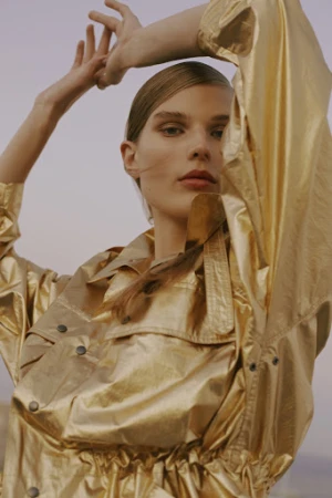 Model wearing a shiny metallic gold jacket with arms raised, makeup by Laure Dansou against an open-air sky backdrop