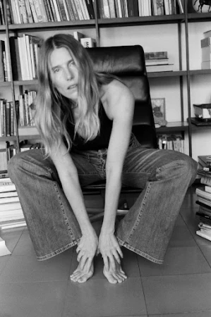 Monochrome portrait of a woman crouched on an office chair with a large bookshelf filled with books in the background.