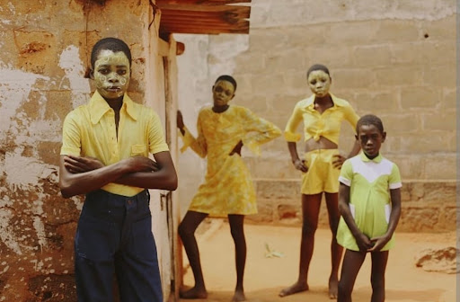 Four young people with painted faces stand outside a building with a dirt ground; they wear yellow- and green-colored garments.
