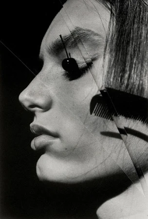 Monochrome profile portrait of a woman with a cherry resting on her eyelid and a comb cutting across her hair.