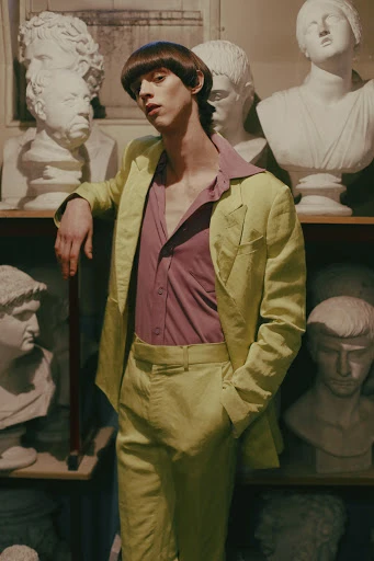 A young adult male with a bowl cut hairstyle poses in a light-green suit, standing indoors among a collection of classic white sculptures.