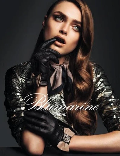 A woman in a snakeskin jacket and black gloves with red-brown wavy hair poses in a studio with medium lighting and a grey backdrop.